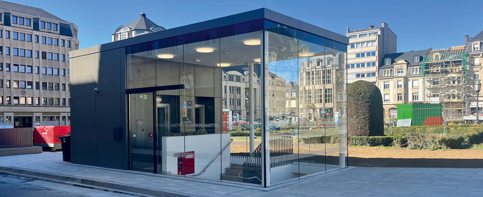 Aussenansicht Parkhauszugang Aussenansicht mit Glassfassade sowie Blechverkleidung vom Parkhauszugang. Im Hintergrund begrünter Platz sowie verschiedene Gebäude.